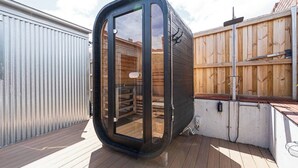 Un sauna extérieur moderne et fermé, doté d’une porte en verre, d’une terrasse en bois et d’une clôture en bois en arrière-plan.