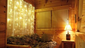 Une chambre confortable dans un chalet, avec un intérieur en bois, un lit recouvert d’une literie à motifs, une petite table avec une lampe et une fenêtre équipée de stores.