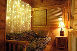 Ein gemütliches Blockhauskabinenzimmer mit Holzvertäfelung, einem Bett mit gemusterten Betttextilien, einem kleinen Tisch mit einer Lampe und einem Fenster mit Jalousien.