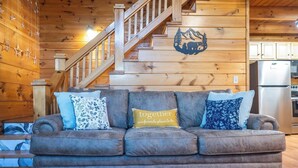 L’intérieur d’un chalet confortable, avec des murs en bois, un escalier en bois, un canapé agrémenté de coussins décoratifs et un coin cuisine.