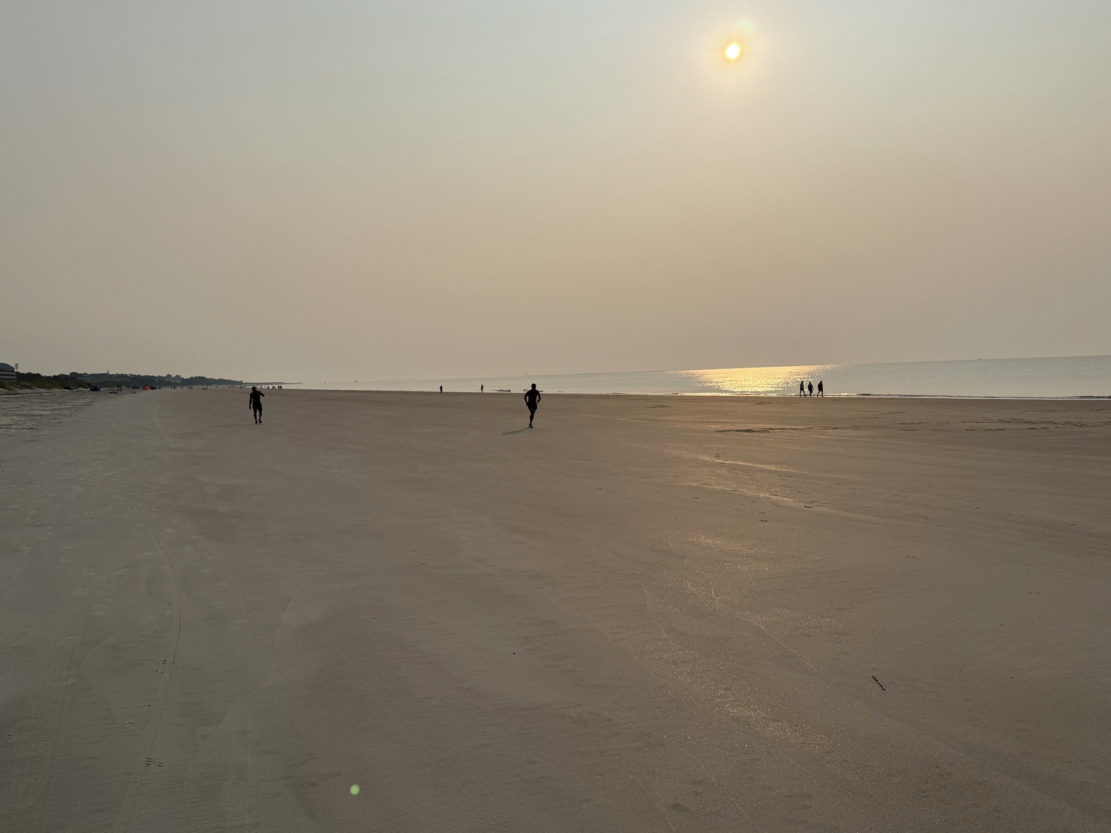 Une vaste plage ouverte, avec quelques personnes qui s’y promènent, le soleil bas à l’horizon et une mer calme en arrière-plan.
