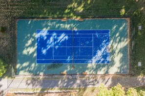 Eine Vogelperspektive zeigt einen Tennisplatz mit umgebender Bepflanzung und einem gepflasterten Weg.