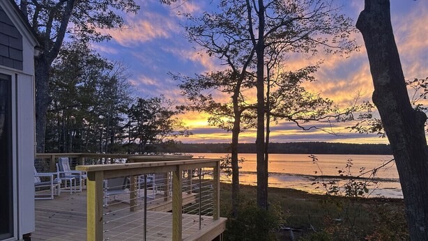Eine Holzterrasse mit Stühlen mit Blick auf einen See bei Sonnenuntergang.