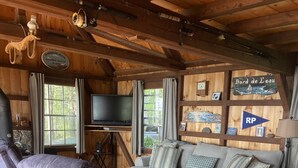 Ein gemütliches Blockhaus-Interieur mit Holzbalkendecke, einer Couch, zwei Sesseln, einem Fernseher und einem Schild mit der Aufschrift 'Bord de L'eau'.