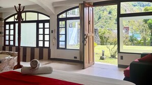 Ein geräumiges Schlafzimmer mit einem großen Bett, einem Holz-Kleiderständer und Blick auf einen üppigen Garten durch große Fenster.