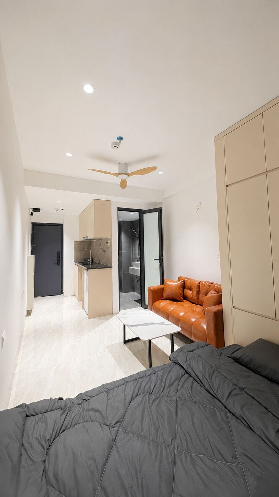 Quarto moderno com cama, sofá, ventilador de teto e uma cozinha compacta ao fundo.