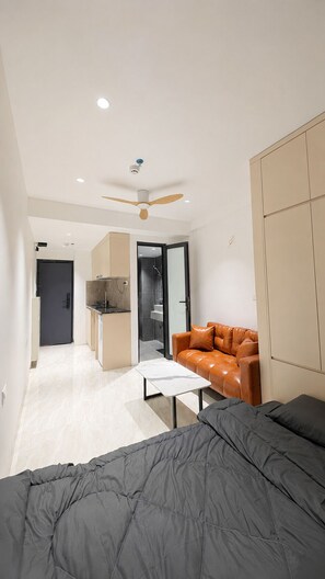 A modern bedroom with a bed, a sofa, a ceiling fan, and a kitchenette in the background. - ERA Trung Liet SIGNATURE (Hanoi)