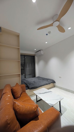A modern living room with a brown leather sofa, a small white coffee table, a ceiling fan, and a bed with a grey duvet.