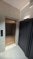 Um elevador com visor digital, porta com placa de identificação e luz de emergência na parede.