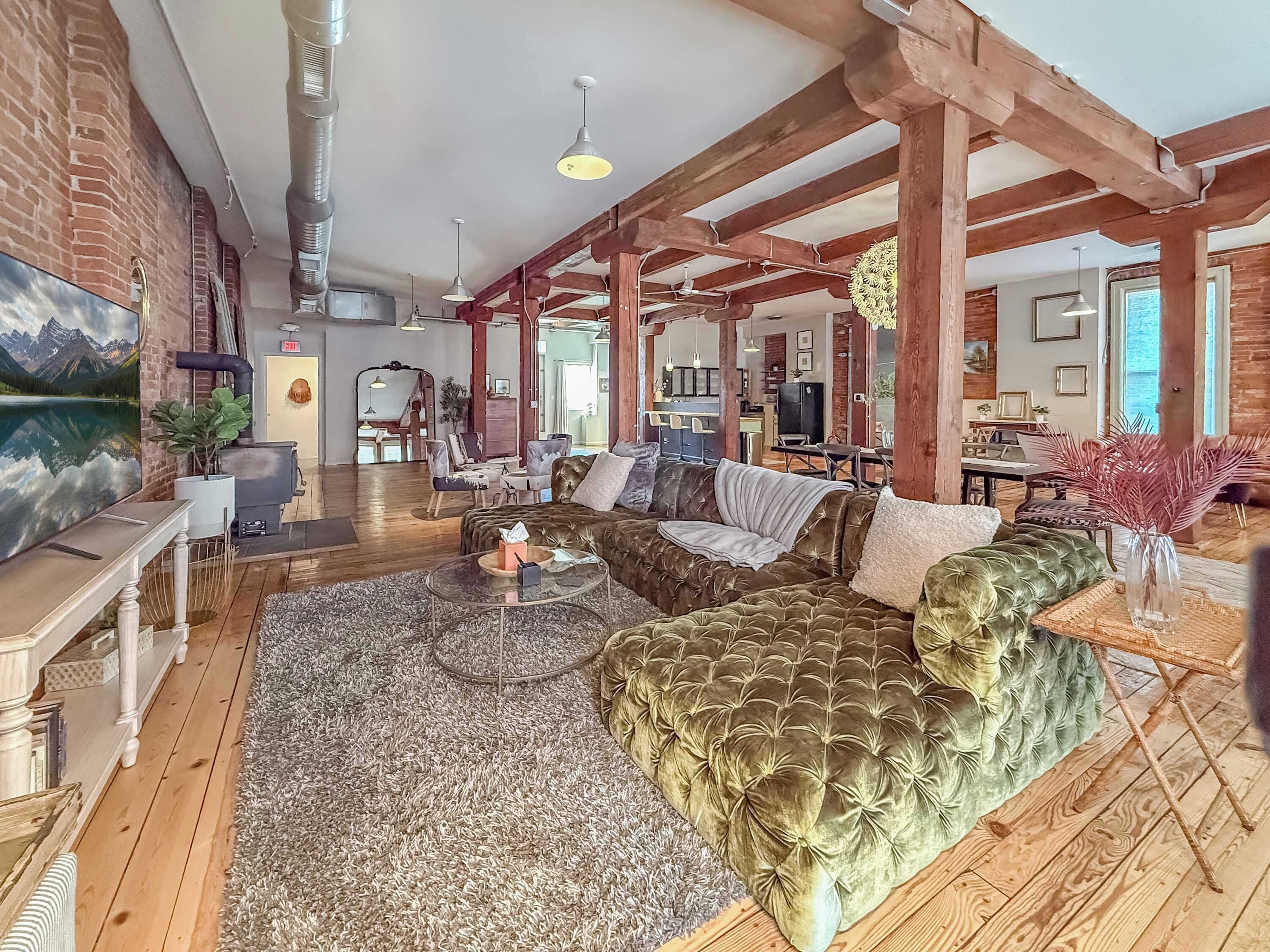A spacious living area with a large sectional sofa, a coffee table, and a fireplace. The room features exposed wooden beams, a brick wall, and a large window.