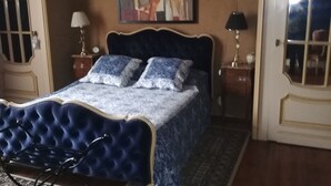 A bedroom with a tufted headboard, a patterned bedspread, two bedside lamps, a rug, and a painting above the bed. - THE LARGE COTTAGE AT CHÂTEAU SAINTE MARIE NEAR THE MARAIS POITEVIN (Fors)