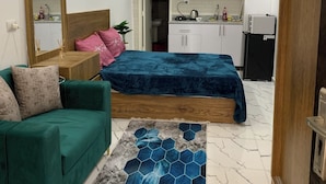 Quarto moderno com sofá verde, cama com colcha azul, tapete hexagonal estampado e cozinha com armários brancos e micro-ondas.