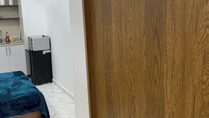 Quarto com porta de madeira, cama com cobre azul, pequena cozinha e chão de azulejos.