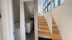 A modern interior with a wooden staircase leading to a second floor, a laundry area with washing machines, and large windows providing natural light.
