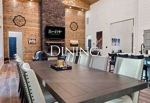 Ein Essbereich mit einem langen Tisch, Stühlen, einem Kamin und einem Fernseher, der 'Trickle Vacation Rentals' anzeigt.