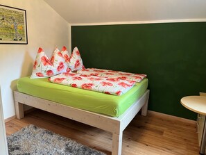 Un lit simple avec une literie verte et des coussins Ă motifs floraux, une petite table ronde et un mur vert.