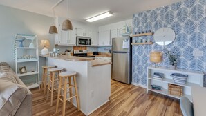 A modern kitchen with white cabinets, a central island with stools, a microwave, and a refrigerator. There is a wooden floor, a white sofa, and a patterned wall. - Winds V D1 - Oceanfront Pet Friendly Condo with Pool and Close to Boardwalk (Carolina Beach)