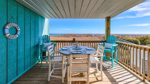 A wooden deck with a table and chairs, a lifebuoy sign, and a view of the beach. - Winds V D1 - Oceanfront Pet Friendly Condo with Pool and Close to Boardwalk (Carolina Beach)