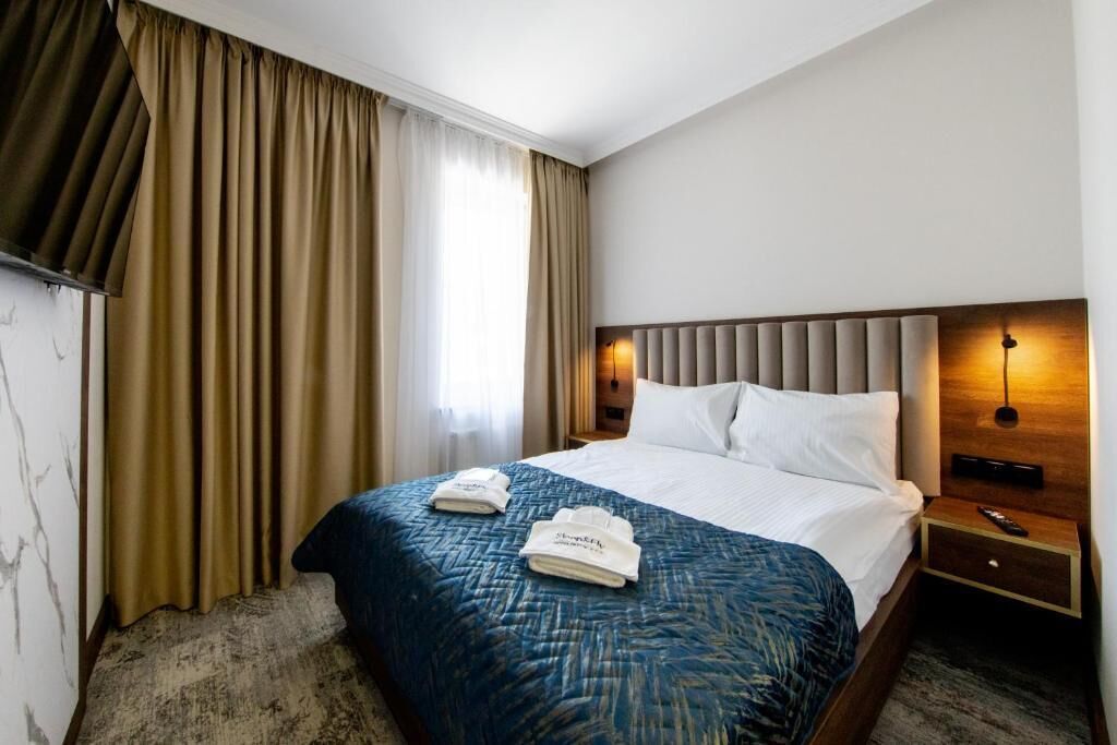 Une chambre d’hôtel avec un grand lit, des rideaux beiges, une tête de lit en bois, une table de chevet et une lampe fixée au mur.