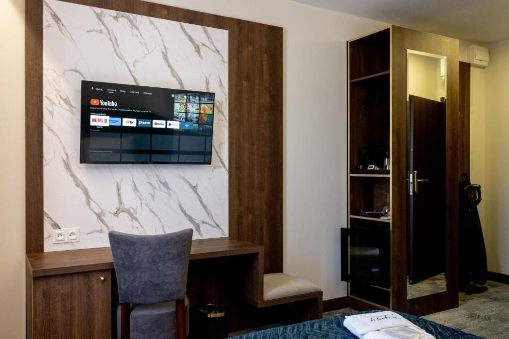 Ein Hotelzimmer mit Flachbildfernseher an einer Marmorwand, einem Holzschrank, einem grauen Sessel und einem Bett mit blauer Bettdecke.