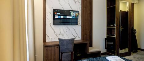 A hotel room with a bed, a desk with a chair, a TV mounted on a marble wall, and a wardrobe.