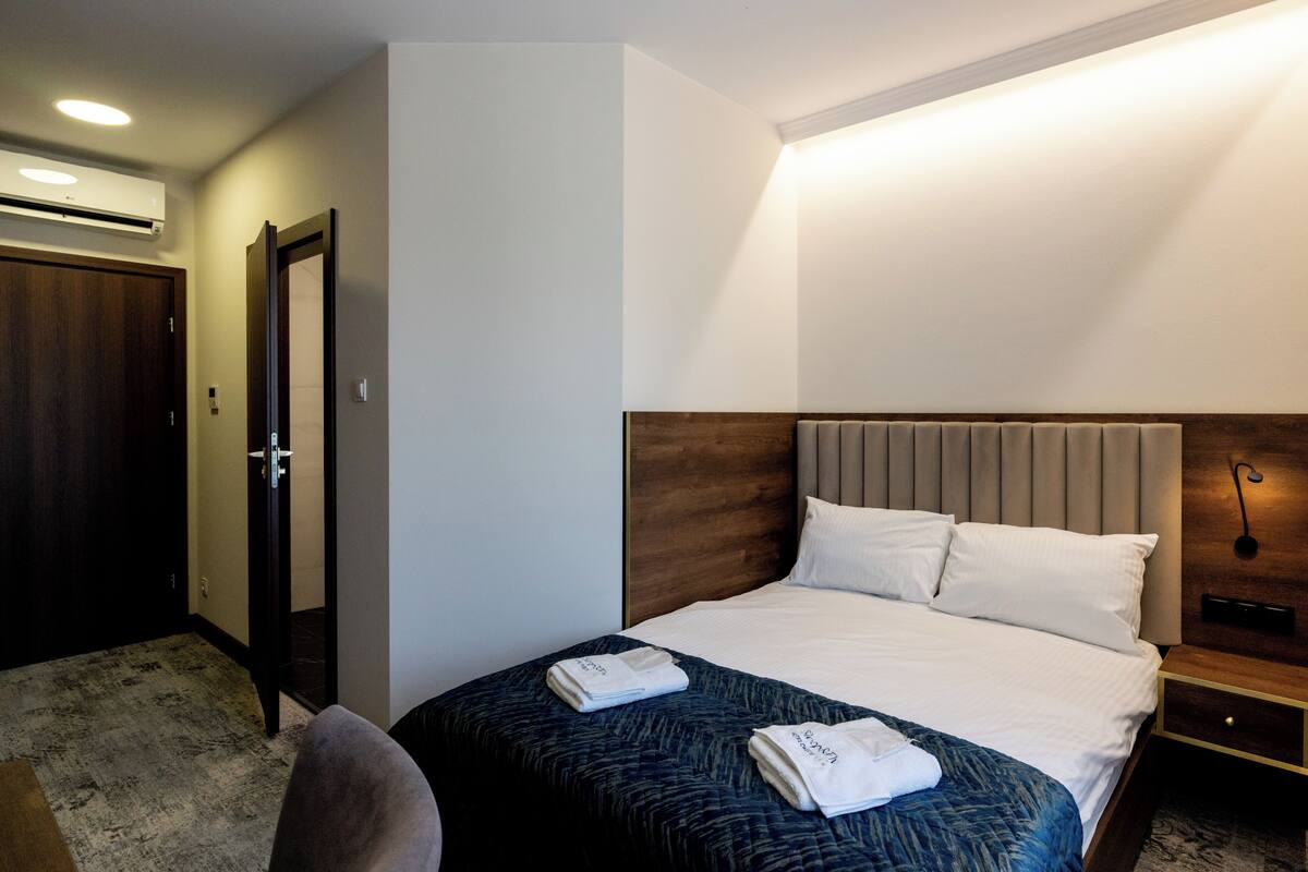 Habitación de hotel con cama, mesita de noche, silla, puerta y equipo de aire acondicionado.