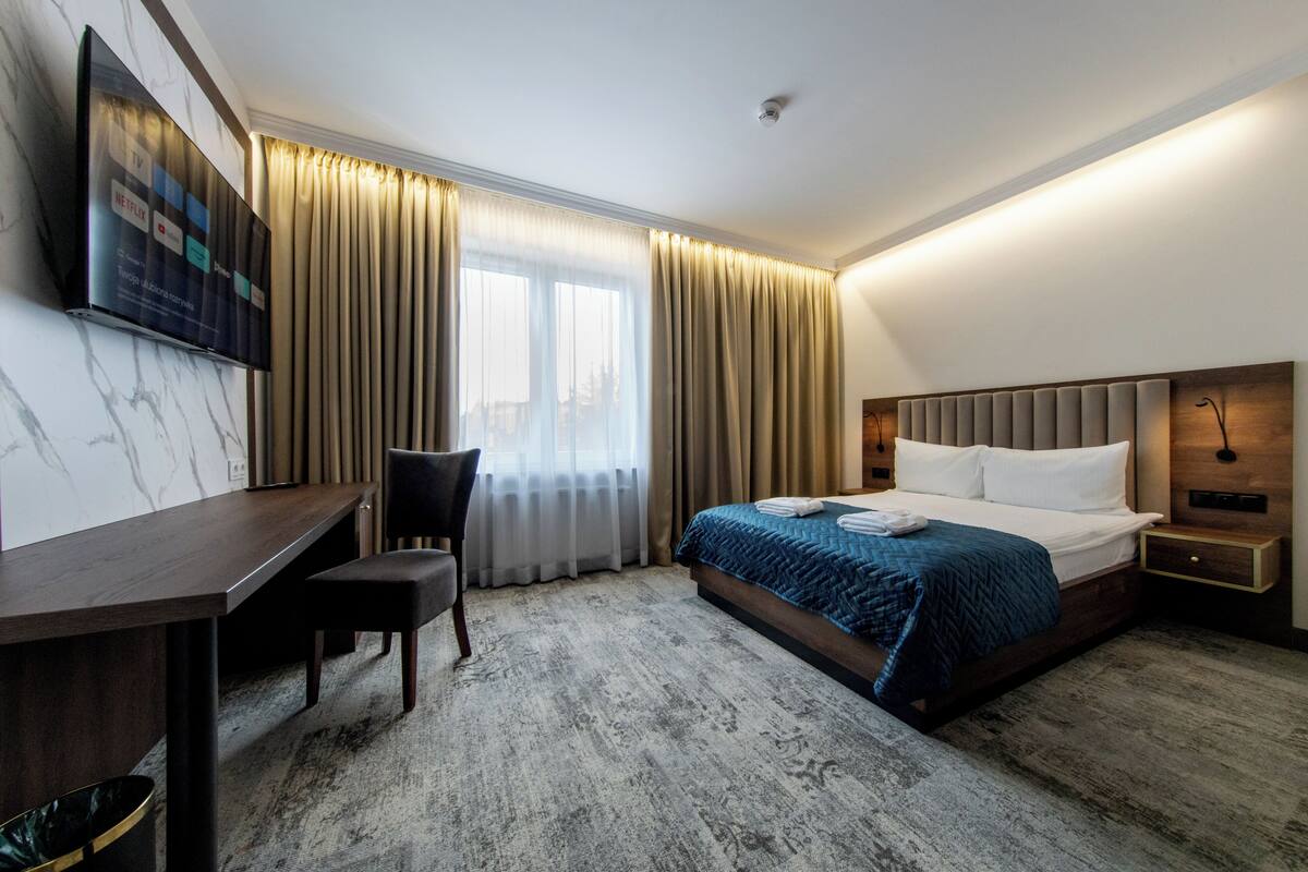 Habitación de hotel con cama, escritorio, silla, televisor montado en la pared y ventana con cortinas.