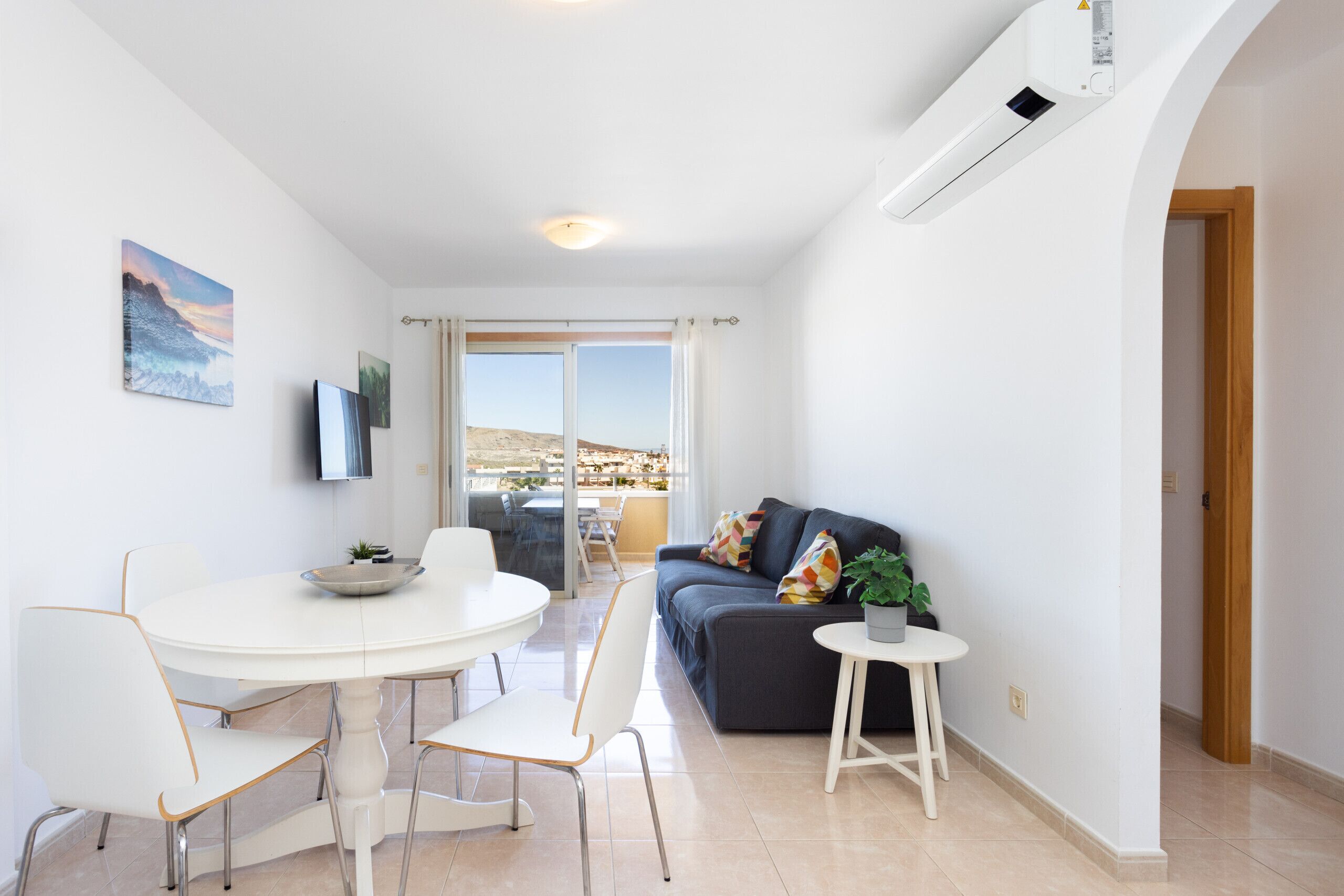 A modern living room with a round dining table, white chairs, a dark sofa, and a small side table with a plant.