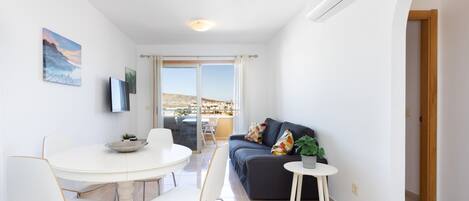 A modern living room with a round dining table, white chairs, a dark sofa, and a small side table with a plant.