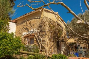 Un bâtiment rustique en pierre, avec un toit de tuiles, entouré de verdure et surplombé d’un ciel bleu clair.