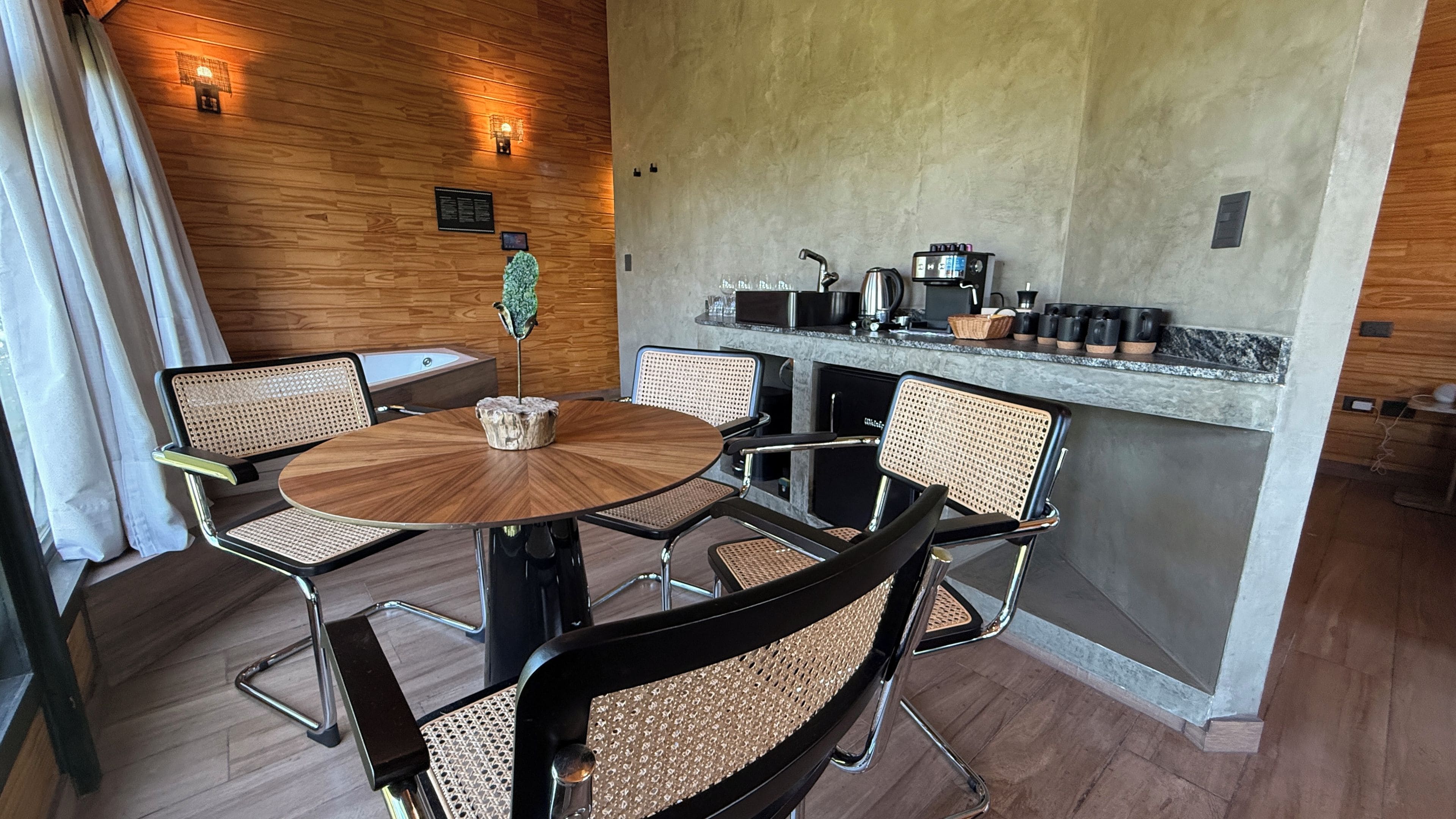 A modern interior with a round wooden table surrounded by six chairs with woven seats, a small cactus plant, and a kitchen area with a coffee machine and various appliances.