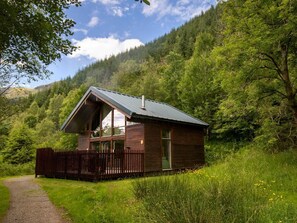 A cabin with a sloped roof, large windows, and a wooden exterior, surrounded by lush greenery and a forested hillside. - Strathyre Classic Golden Oak, Strathyre, Ben Ledi (Callander)