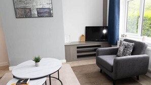 Ein modernes Wohnzimmer mit grauem Sofa, einem kleinen runden Tisch mit Pflanze, einem Flachbildfernseher auf einem Holzgestell und einem Fenster mit Blick auf Grünflächen.