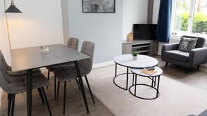 Ein modernes Wohnzimmer mit Esstisch, zwei Sesseln, einem Couchtisch, einem Fernseher und einem fenster mit Vorhängen.