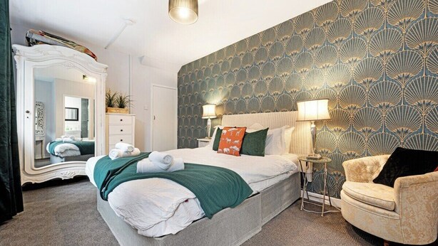 A hotel room with a large bed, a vanity with a mirror, a chair, and a patterned wallpaper. - Regency by the Sea @ Blackpool (Blackpool)