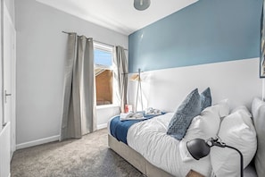 A bedroom with a bed, a lamp, a window with curtains, and a view of a building outside. - StayMinorca @ Bolton (Bolton)