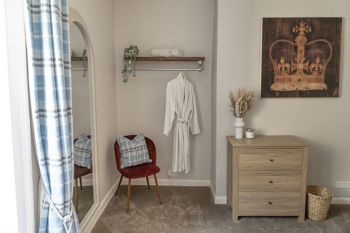 Ein Badezimmer mit einem Holzkosmetikschrank, einem roten Stuhl, einem Handtuchhalter mit Bademantel, einem Spiegel und einem wandmontierten Ablagebereich mit einer Pflanze.