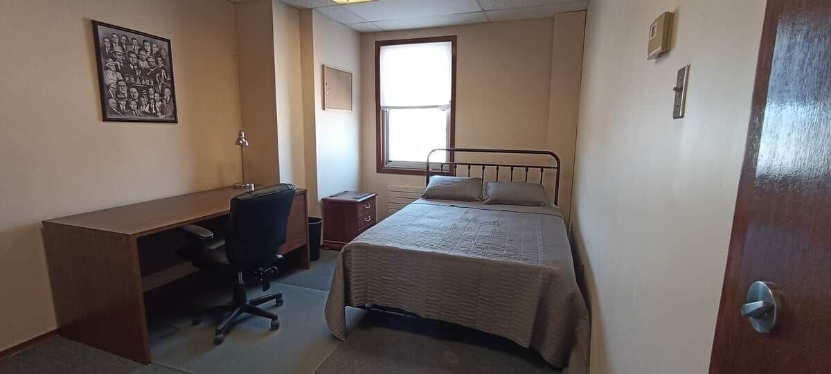 Ein Schlafzimmer mit einem Bett, einem Schreibtisch, einem Stuhl, einem Fenster und einer Tür.