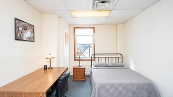 Um quarto pequeno e bem arrumado, com uma cama, uma escrivaninha, uma cadeira e uma mesinha de cabeceira.