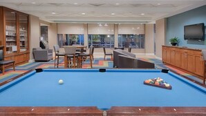 A pool table with a blue felt surface, surrounded by chairs and a bookshelf.