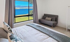 A bedroom with a bed, a sofa, a window with a view of the sea, and curtains. - Absolute Lake Front Apartment (East Jindabyne)