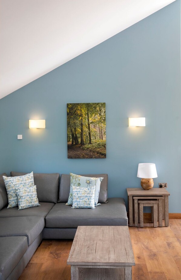 Ein Wohnzimmer mit einem grauen Ecksofa, zwei Wandleuchten, einem Holztisch und einem Gemälde eines Waldes.