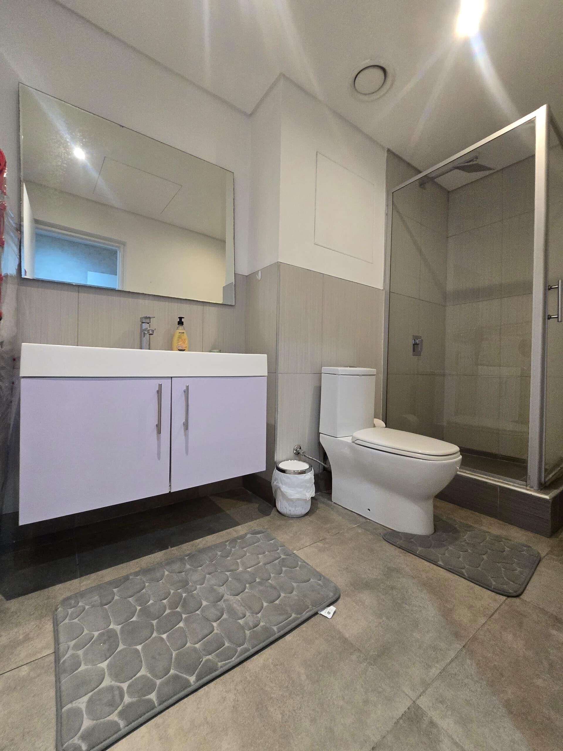 Een moderne badkamer met een wit badkamermeubel, een toilet, een douchecabine van glas en een badmat met steenpatroon.