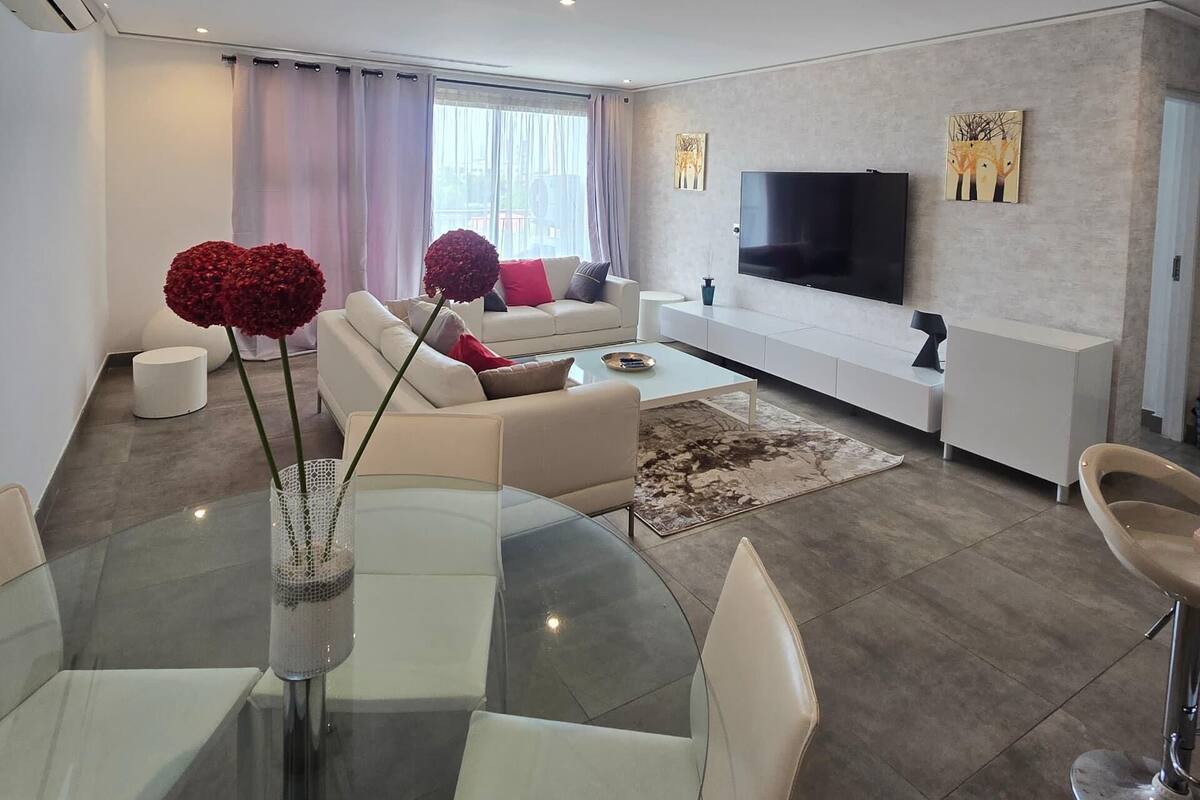 A modern living room with a glass dining table, white sofas, a flat-screen TV, and a large window with curtains.