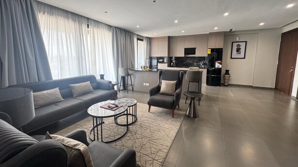A modern living room with a sofa, armchairs, a coffee table, and a kitchen in the background. - Bravia Residence by Cozy (Accra)