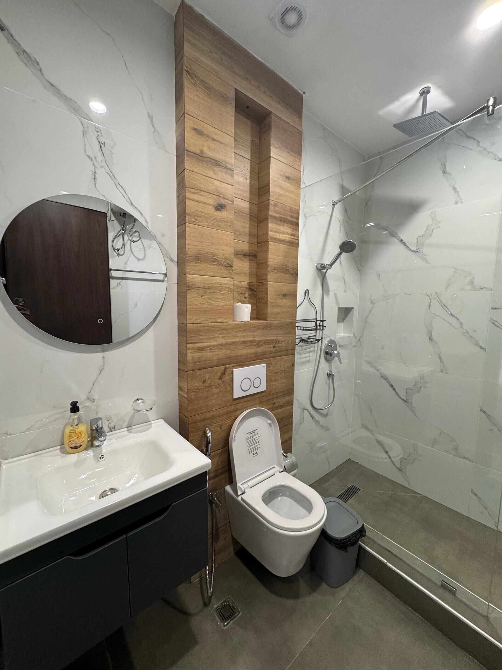 A modern bathroom with a wooden vanity, a round mirror, a toilet, and a glass-enclosed shower area.