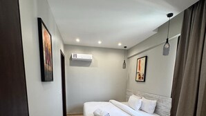 A modern hotel room with a large bed, an air conditioning unit, a small bedside table, and a painting on the wall. - Bravia Residence by Cozy (Accra)