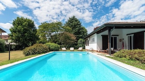 Ein rechteckiger Swimmingpool mit klarem, blauem Wasser, umgeben von Grünpflanzen und einem Gebäude mit einem Ziegeldach.