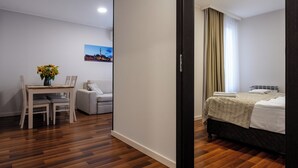 A hotel room with a wooden floor, a bed with a white duvet, a small table with a vase of flowers, a grey sofa, and a picture on the wall. - Wesoła Apartamenty (Wólka Kosowska)