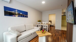 A modern living room with a sofa, a wooden coffee table with fruit, a dining area with chairs, and a TV mounted on the wall. - Wesoła Apartamenty (Wólka Kosowska)
